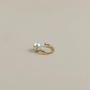 May include: A gold-colored ear cuff featuring a single, round, white pearl. The cuff curves around the ear, with a small ball at the end of the curve. The jewelry is set against a neutral background.
