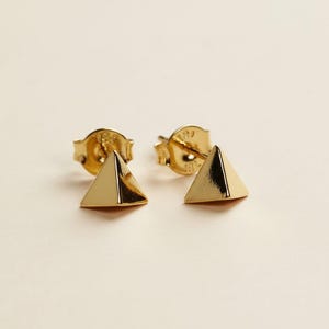 May include: A pair of gold-colored pyramid stud earrings. The earrings have a simple, geometric design with a polished finish. The posts and backs are also gold-colored. The earrings are displayed on a light beige background.