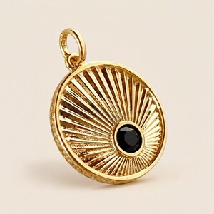 May include: A circular gold pendant featuring a sunburst pattern of radiating lines and a single round black gemstone set off-center. The pendant includes a small gold jump ring at the top for attachment to a chain or cord.