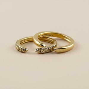 May include: Two gold-toned open rings with a polished finish. Each ring features a tapered end encrusted with small, clear, sparkling stones. The rings are positioned overlapping each other, showcasing a smooth band on one side and textured detail.