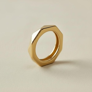 Puede incluir: Un anillo de tono dorado pulido que presenta un diseño exterior geométrico y hexagonal de múltiples facetas. El anillo tiene una superficie metálica lisa y reflectante con bordes afilados y limpios que crean una apariencia angular y estructurada.