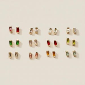 May include: A collection of twelve pairs of rectangular baguette-cut stud earrings set in gold-toned metal. Each pair features a different colored gemstone, including red, clear, green, pink, orange, and blue hues, arranged in a grid pattern.