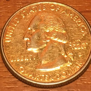 Może przedstawiać: Złota moneta ćwierćdolara amerykańskiego z wizerunkiem George'a Washingtona na awersie. Na monecie widnieje napis "United States of America" wokół krawędzi oraz "Liberty" i "Quarter Dollar" pod wizerunkiem. Napis "In God We Trust" jest widoczny po prawej stronie monety.