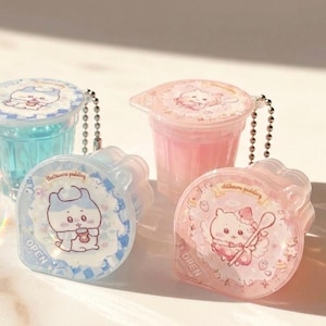 May include: Four small, clear plastic containers with colorful contents and decorative lids. Two containers are blue, and two are pink. Each lid features a cartoon character and the text "OPEN". The containers are attached to silver ball chains.
