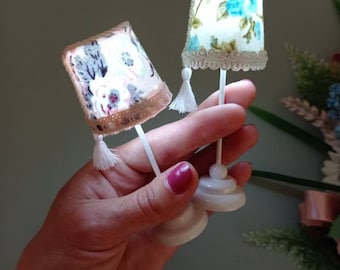 Può includere: Due lampade da tavolo in miniatura con basi bianche e paralumi in tessuto. I paralumi hanno un motivo floreale e un bordo bianco. Le lampade hanno nappine bianche.