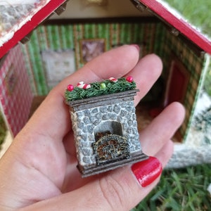 Può includere: Un camino in pietra in miniatura con una griglia in metallo nero e una ghirlanda verde con bacche rosse in cima. Il camino è fatto di pietra grigia e ha una base in legno marrone.