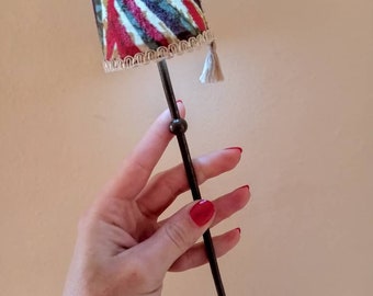 Può includere: Una lampada da tavolo in miniatura con un supporto in metallo nero e un paralume in tessuto colorato. Il paralume ha un fiocco e un bordo marrone.
