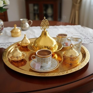 May include: A golden serving tray with ornate detailing holds a cup of coffee, a covered dish, and a small dish with a lid. Two glasses of water and a small dish with a piece of Turkish delight are also on the tray. The tray is on a dark brown table.