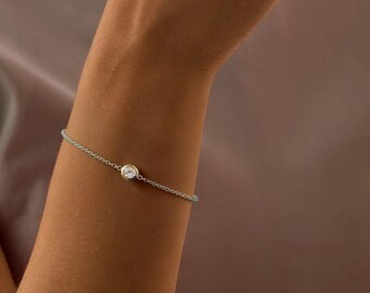 May include: A delicate silver bracelet featuring a single, round, clear gemstone. The bracelet is a simple chain design, and is worn on a person's wrist. The background is a soft, neutral color.