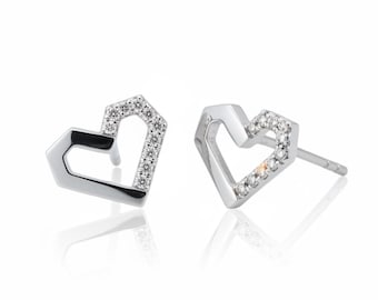 May include: A pair of silver-colored stud earrings in a heart shape. One side of the heart is open, while the other side is encrusted with small, clear gemstones. The earrings are displayed on a white background.