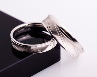 May include: Two silver wedding bands with a textured, diagonal line design. The bands are on a black surface.