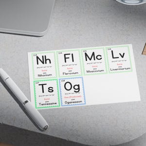 May include: A white card featuring a periodic table of elements, including Nihonium, Flerovium, Moscovium, Livermorium, Tennessine, and Oganesson. The elements are in a grid with their symbols, atomic numbers, and states of matter. A pen rests on the table.