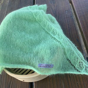 May include: A light green, hand-knitted shawl with a delicate, lacy edge. The shawl is made of a soft, fuzzy yarn and is resting on a wooden surface. A small tag reads "Hand-Knitted by Ruchfield Knits."