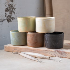 Puede incluir: Cinco tazas de cerámica de diferentes colores, incluyendo verde, amarillo, marrón y negro, apiladas sobre una superficie de madera. Dos herramientas de cerámica de madera están en primer plano.