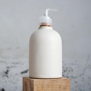 Könnte beinhalten: Ein cremefarbener Seifenspender aus Keramik mit strukturierter Oberfläche. Er hat eine transparente Kunststoffpumpe und ein Korkdetail am Hals. Der Spender steht auf einem Holzblock vor neutralem Hintergrund.