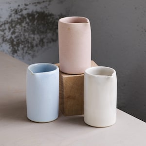 Può includere: Tre vasi in ceramica dai colori pastello: azzurro, rosa pallido e bianco. Ogni vaso ha una forma cilindrica con un bordo leggermente irregolare. I vasi sono disposti su un blocco di legno e una superficie chiara.