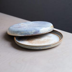 Puede incluir: Dos platos de cerámica, uno apilado sobre el otro. Los platos tienen un esmalte azul claro y marrón con una apariencia rústica y hecha a mano. Los platos son redondos con un borde ligeramente elevado.
