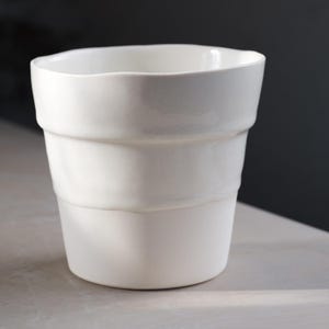Puede incluir: Una taza de cerámica blanca con un diseño único en capas. La taza tiene un borde ligeramente irregular y un acabado liso y mate. El diseño presenta tres bandas horizontales que le dan a la taza una apariencia texturizada. Adecuada para bebidas.