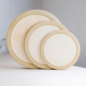 May include: A set of three circular ceramic plates of varying sizes, each featuring a smooth, off-white center and a wide, textured, light beige rim. The plates are arranged in a cascading stack, showcasing their round shape and contrasting tones.