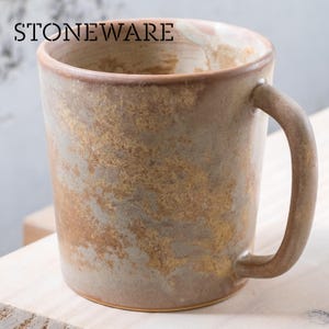 Puede incluir: Una taza de gres con un acabado rústico y texturizado. La taza tiene forma cilíndrica con un asa curvada. El exterior presenta una mezcla de tonos marrones, beige y grises, con la palabra "STONEWARE" impresa en la parte superior. Adecuada para café, té u otras bebidas.