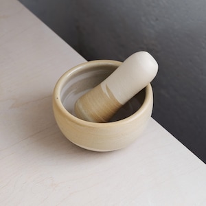 May include: A ceramic mortar and pestle. The mortar is light beige and round. The pestle is cylindrical, with a light beige top and a darker beige band. This set is designed for grinding herbs and spices, ideal for the kitchen.