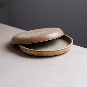May include: A handmade, ceramic serving dish with a lid. The dish is a warm, earthy tone with a slightly textured surface. The lid is partially open, revealing the interior of the dish. The overall design is simple and elegant, suitable for various uses.