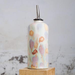 Può includere: Una bottiglia per olio in ceramica con beccuccio in acciaio inossidabile. La bottiglia è cilindrica con base bianca e motivi astratti colorati nelle tonalità dell'arancione, del viola e del verde. È appoggiata su un blocco di legno.