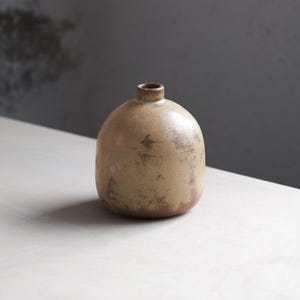 Puede incluir: Un jarrón pequeño, redondo, de cerámica con un esmalte marrón claro y un acabado moteado. El jarrón tiene una abertura estrecha en la parte superior.