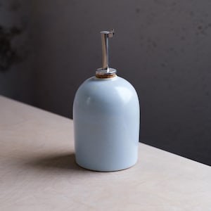 May include: A light blue ceramic oil bottle with a stainless steel spout and a cork stopper. The bottle has a rounded shape and is sitting on a light-colored surface. The spout is angled and has a small lever.