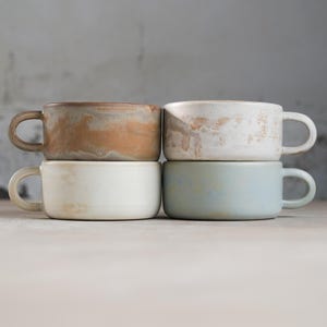Pode incluir: Quatro canecas de cerâmica empilhadas em dois pares. As canecas são em vários tons de marrom, branco e azul com um esmalte salpicado.