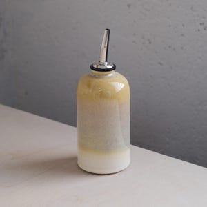 Può includere: Una bottiglia per olio in ceramica con beccuccio in acciaio inossidabile. La bottiglia ha una sfumatura di colore, che va dal bianco alla base al beige e giallo nella parte superiore. Il beccuccio è argentato e la bottiglia è cilindrica.