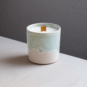 Puede incluir: Un recipiente cilíndrico de cerámica para velas con un esmalte de dos tonos, que presenta una sección superior de color verde salvia suave que transiciona hacia una base de color blanco crema. La vela contiene cera blanca y una mecha de madera.