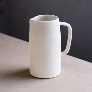 Può includere: Una brocca alta in ceramica bianco sporco con manico. La brocca ha una superficie leggermente ruvida e un design semplice ed elegante. Il bordo interno è di una tonalità più chiara. Adatta per servire acqua, succo o altre bevande.