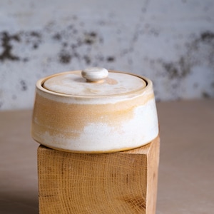 Op de afbeelding: Een kleine, ronde keramische pot met een deksel. De pot is wit met een band van bruin en crèmekleurig glazuur in het midden. Het deksel heeft een kleine, ronde knop. De pot staat op een houten blok.