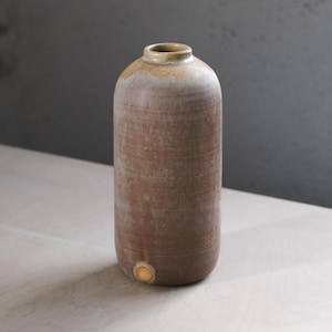 May include: A tall, cylindrical ceramic vase with a light brown glaze and a slightly textured surface. The vase has a rounded bottom and a slightly flared opening.
