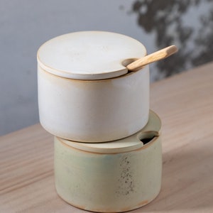 Puede incluir: Dos recipientes de azúcar de cerámica, uno blanco con tapa y una cuchara de madera, apilados sobre un recipiente verde.