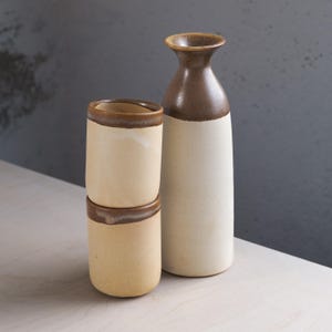 Puede incluir: Un juego de sake de cerámica con una botella alta con la parte superior marrón y dos tazas cilíndricas apiladas. La botella y las tazas tienen una base moteada de color blanquecino con bordes marrones. El juego está diseñado para servir sake u otras bebidas.