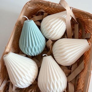 Puede incluir: Una cesta llena de virutas de papel marrón contiene cinco velas con forma de pera. Dos velas son blancas y tres son azul claro. Las velas tienen un diseño texturizado y plisado.