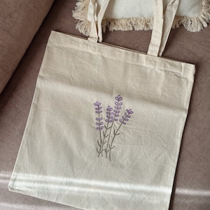 May include: A white canvas tote bag with a simple embroidered design of lavender flowers in shades of purple and green.