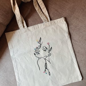May include: A white canvas tote bag with a black line drawing of a person's face with flowers growing from their head. The flowers are embroidered in pink, yellow, blue, and green thread.