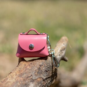 May include: A small pink leather purse keychain with a silver clasp and a silver loop. The keychain is sitting on a piece of weathered wood.