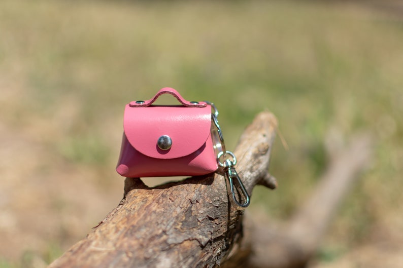 May include: A small pink leather purse keychain with a silver clasp and a silver loop. The keychain is sitting on a piece of weathered wood.