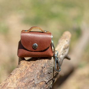 May include: A small brown leather purse with a silver clasp and a silver key chain attached. The purse is sitting on a piece of weathered wood.