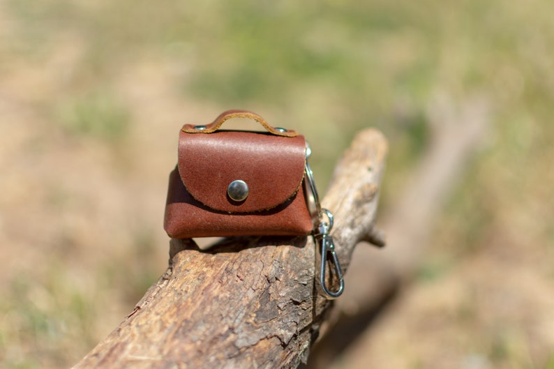 May include: A small brown leather purse with a silver clasp and a silver key chain attached. The purse is sitting on a piece of weathered wood.