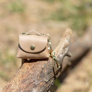 May include: A small, light brown leather pouch with a gold clasp and a gold key chain. The pouch is sitting on a piece of weathered wood.