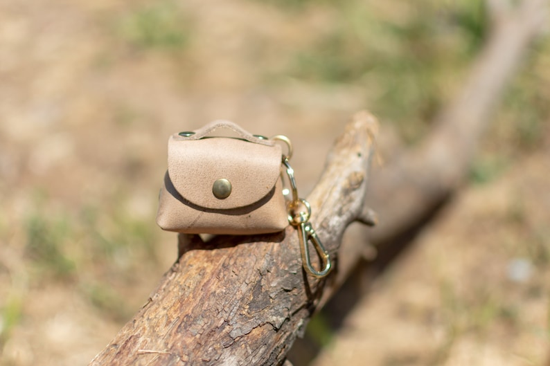 May include: A small, light brown leather pouch with a gold clasp and a gold key chain. The pouch is sitting on a piece of weathered wood.