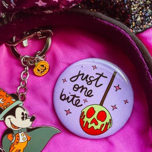 May include: A circular pin featuring a light purple background with the text "just one bite" and a graphic of a red and green skull-shaped candy apple. Beside it is a metal keychain depicting a cartoon character in a cape and a pumpkin charm.