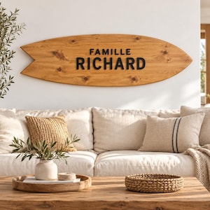 Peut inclure: Une enseigne murale en bois en forme de planche de surf avec une finition en grain de bois brun clair naturel. L'enseigne présente le texte noir FAMILLE RICHARD centré sur le devant. La planche a une pointe et une queue encochée.