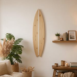 Op de afbeelding: Een wanddecoratie in de vorm van een lichthouten surfboard met een verticale witte streep in het midden. Het surfboard heeft een puntige bovenkant en een gespleten staart, met zichtbare natuurlijke houtnerven en noesten.
