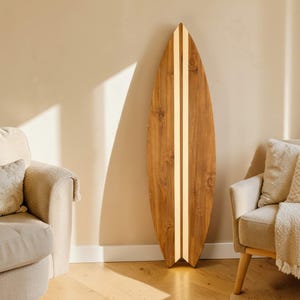 May include: A tall, pointed surfboard made of natural brown wood with a prominent vertical light-coloured stripe running down the centre. The board features a visible wood grain pattern and a split tail design, standing upright against a wall.